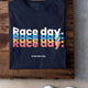 Navy blue t-shirt with 'Race day. Race day. Race day.' text in multicolor on a wooden surface.