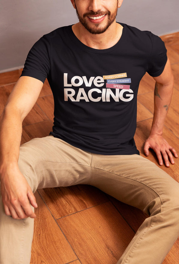 Young man sitting on a wooden floor, wearing a black "Love Racing" t-shirt with bold white text and coloured blocks reading “Every Corner,”“Every Straight,” and “Speed.” Casual, modern look with beige pants, relaxed pose, and a tattoo on his forearm.