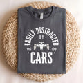 A gray t-shirt with the text 'Easily Distracted - By - Cars' in white. A white cartoon race car is between the words 'By' and 'Cars'. The text is in a circle with a white checkered flag on each side of the circle.