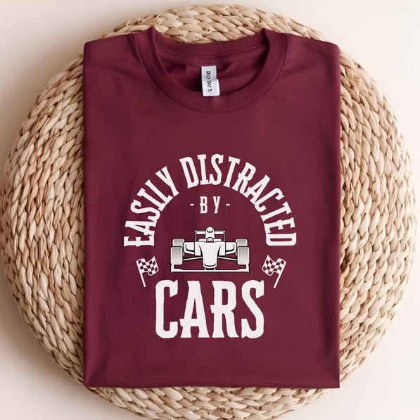 A maroon t-shirt with a white graphic that reads 'Easily Distracted - By - Cars' with a white cartoon race car in the middle of the text.