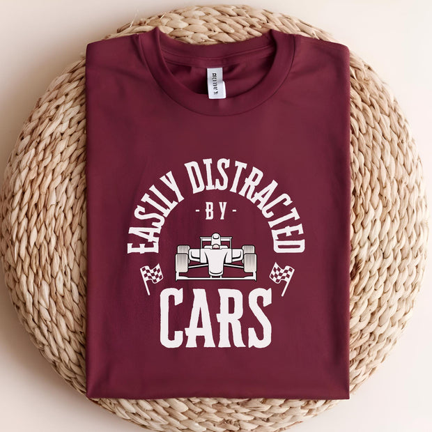 A maroon t-shirt with a white graphic that reads 'Easily Distracted - By - Cars' with a white cartoon race car in the middle of the text.